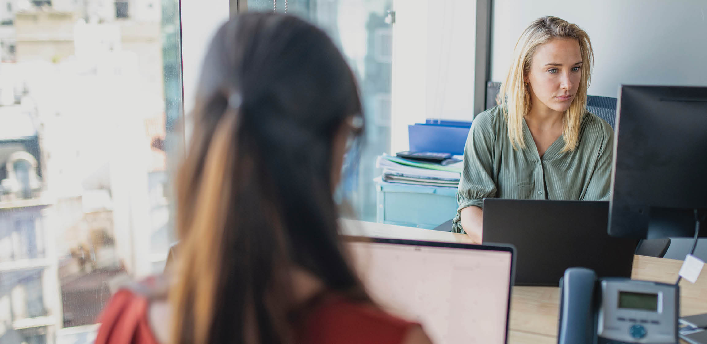Focus on background of blonde and brunette Argentine businesswomen working on laptops while sitting face to face at window desks 