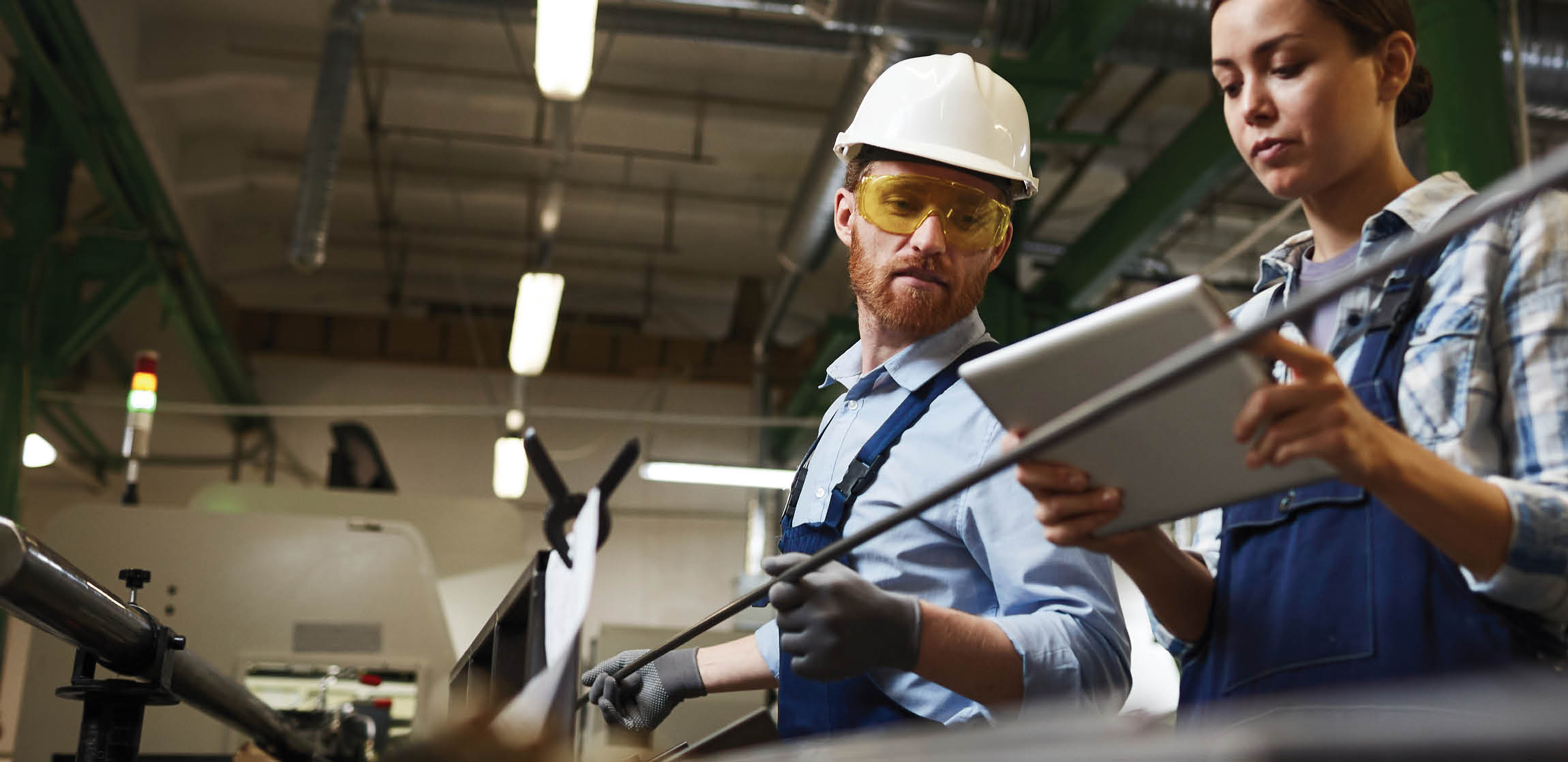 Female foreman in work wear using digital tablet and controlling the work while man working with iron pipes in the plant